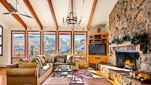 Ein gemütliches Wohnzimmer mit Kamin, großen Fenstern und Blick auf die Berge sowie einer Wand aus Stein.