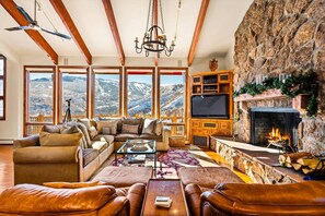 A cozy living room with a fireplace, leather sofas, a large TV, and a scenic mountain view through large windows.