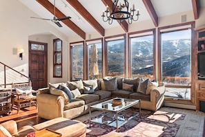 A spacious living room with a large sectional sofa, a glass coffee table, and a view of snow-covered mountains through large windows.