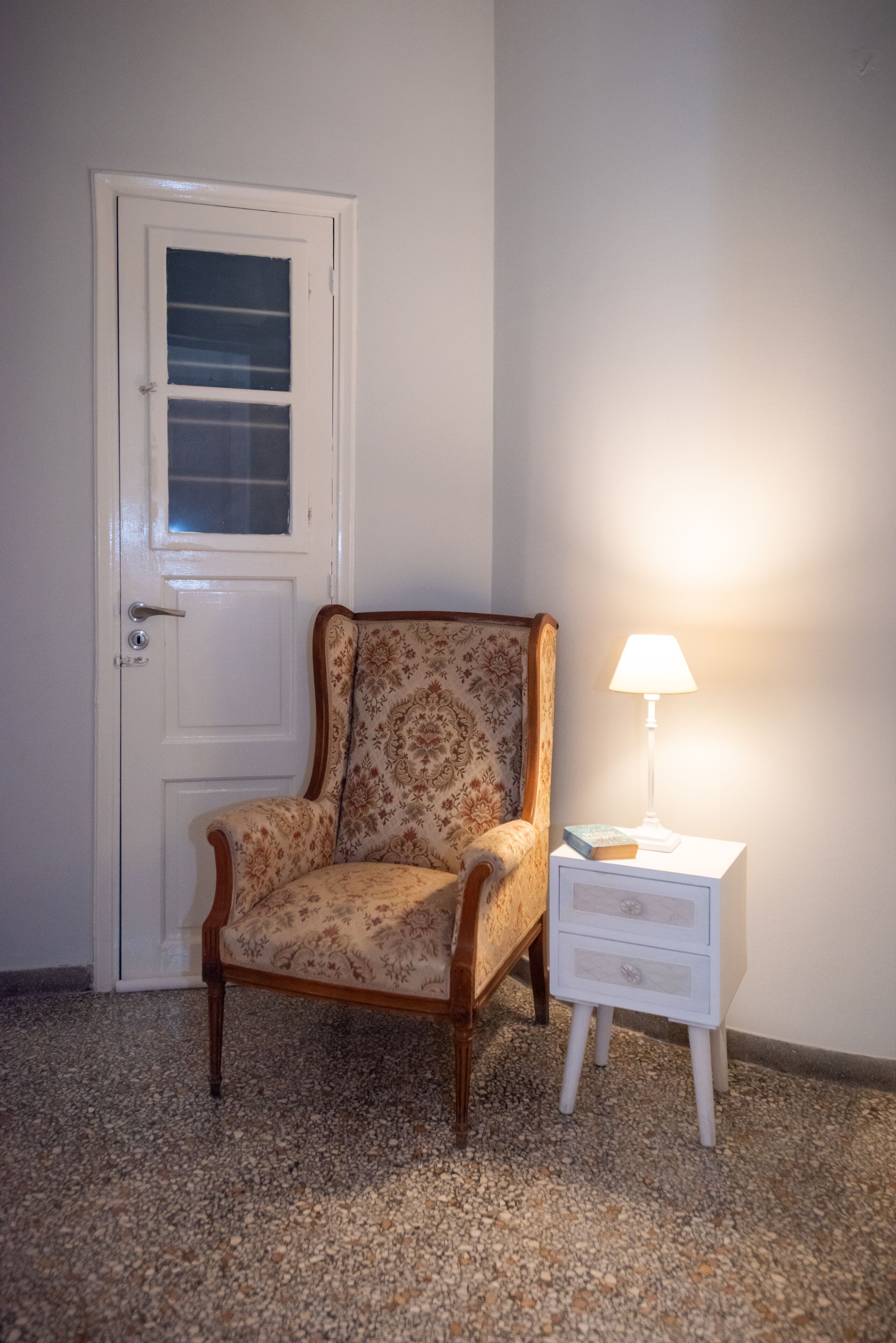 A room with a wooden chair, a white bedside table with a lamp, and a white door with a window.