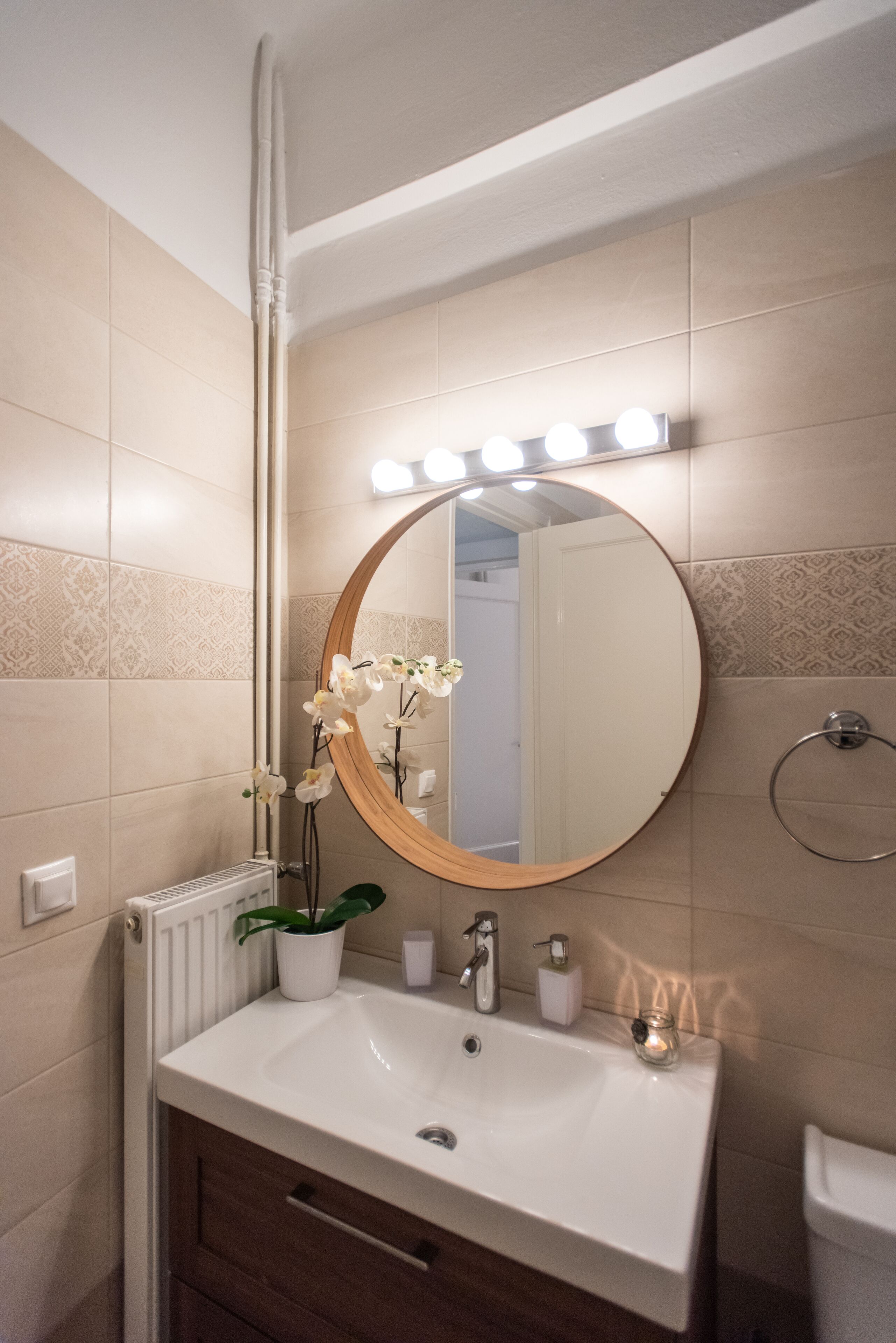 A bathroom with a white sink, a round mirror with lights, a potted orchid, and a towel ring.