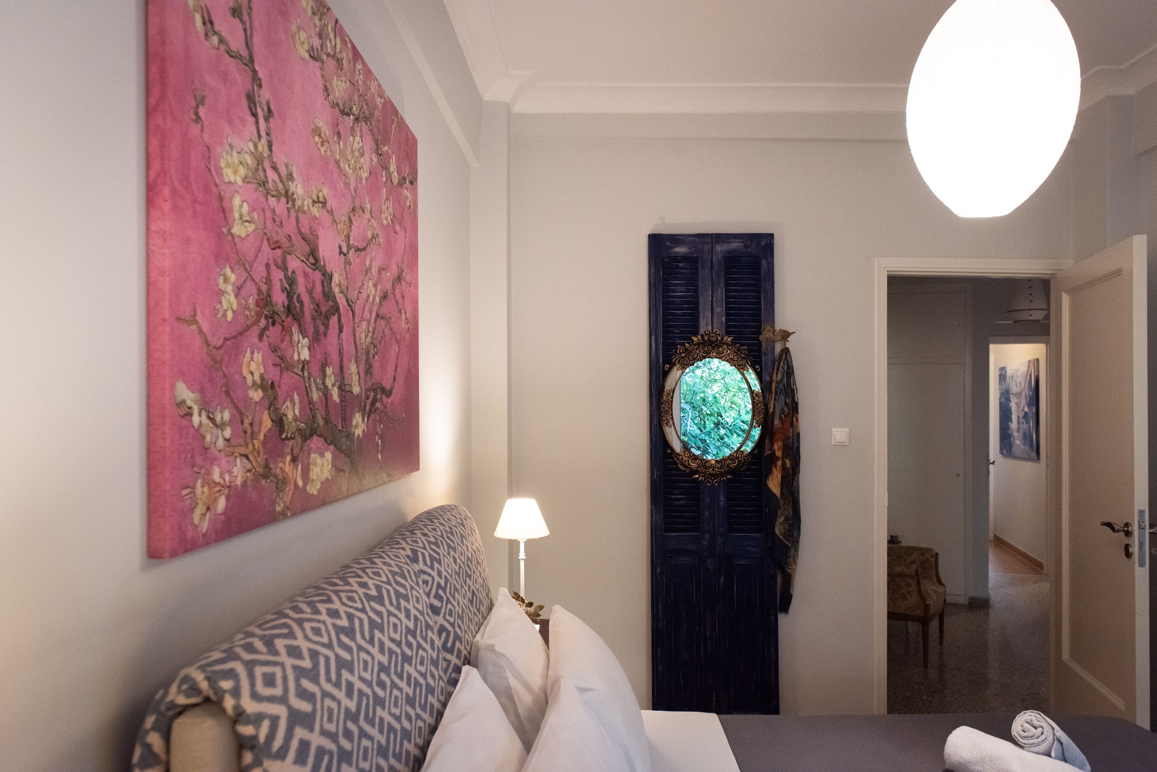Ein Hotelzimmer mit Bett, Nachttisch mit Lampe, großem Blumenbild an der Wand, dunkler Holztür und Flur zu einem anderen Zimmer.