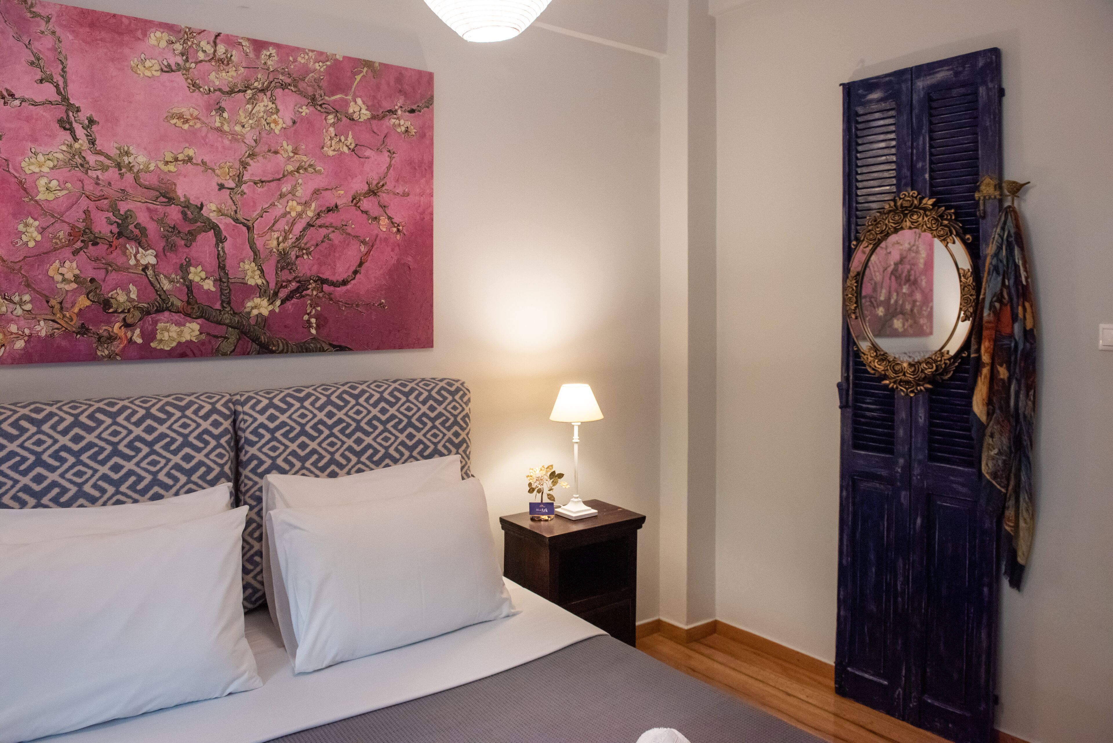 Un dormitorio con cama, almohadas, mesita de noche con lámpara, un cuadro y una puerta con espejo.