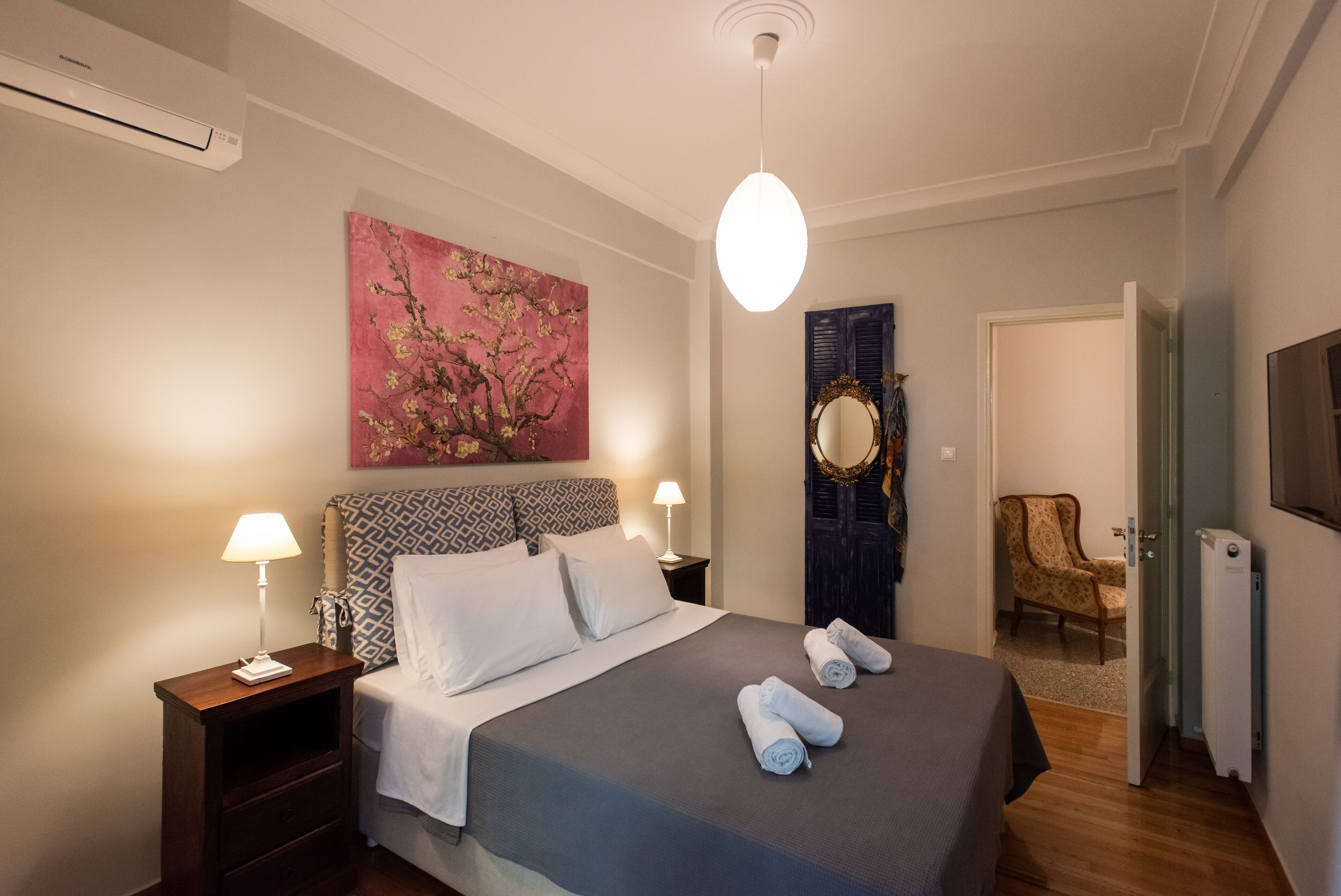 A neatly made bed with pillows and rolled towels, a bedside lamp, a wall painting, a mirror, and a chair in a hotel room.