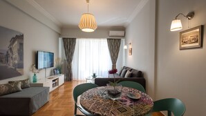 Una sala de estar moderna con un sofá, una mesa de comedor con sillas, un televisor y un amplio ventanal con cortinas.