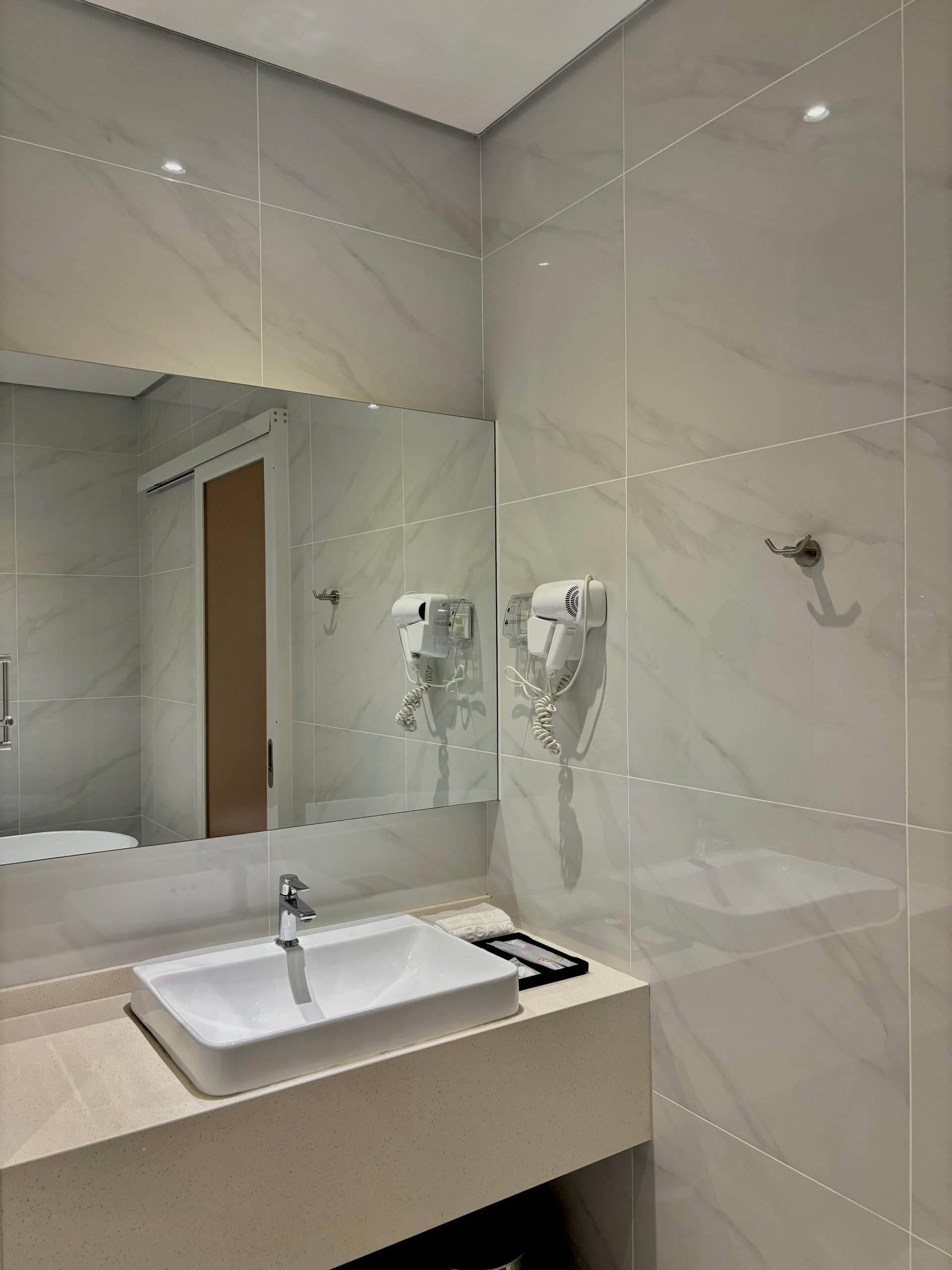 A modern bathroom with a large mirror, a white sink, and two hair dryers mounted on the wall.
