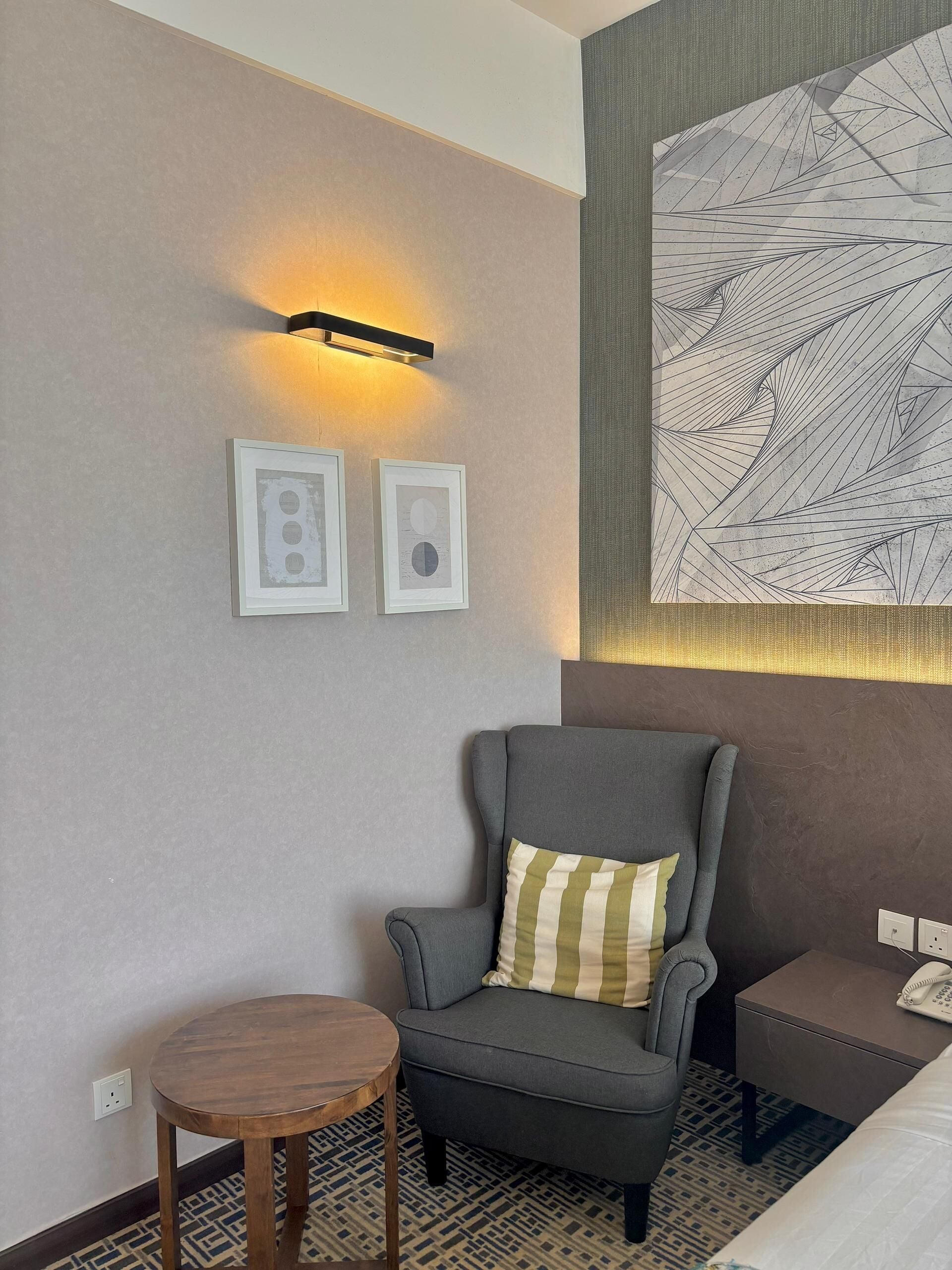 Una habitación de hotel moderna con un sillón gris, una pequeña mesa redonda de madera y un cuadro abstracto grande en la pared.
