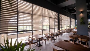 Um espaço amplo e interno, com grandes janelas, mesas e cadeiras de madeira, e plantas em vasos.