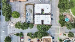 An aerial view of a parking lot with multiple cars, a building with a flat roof, and surrounding greenery. - Downtown Studio by Toyota Center/Minute Maid/ George R Brown (Houston)