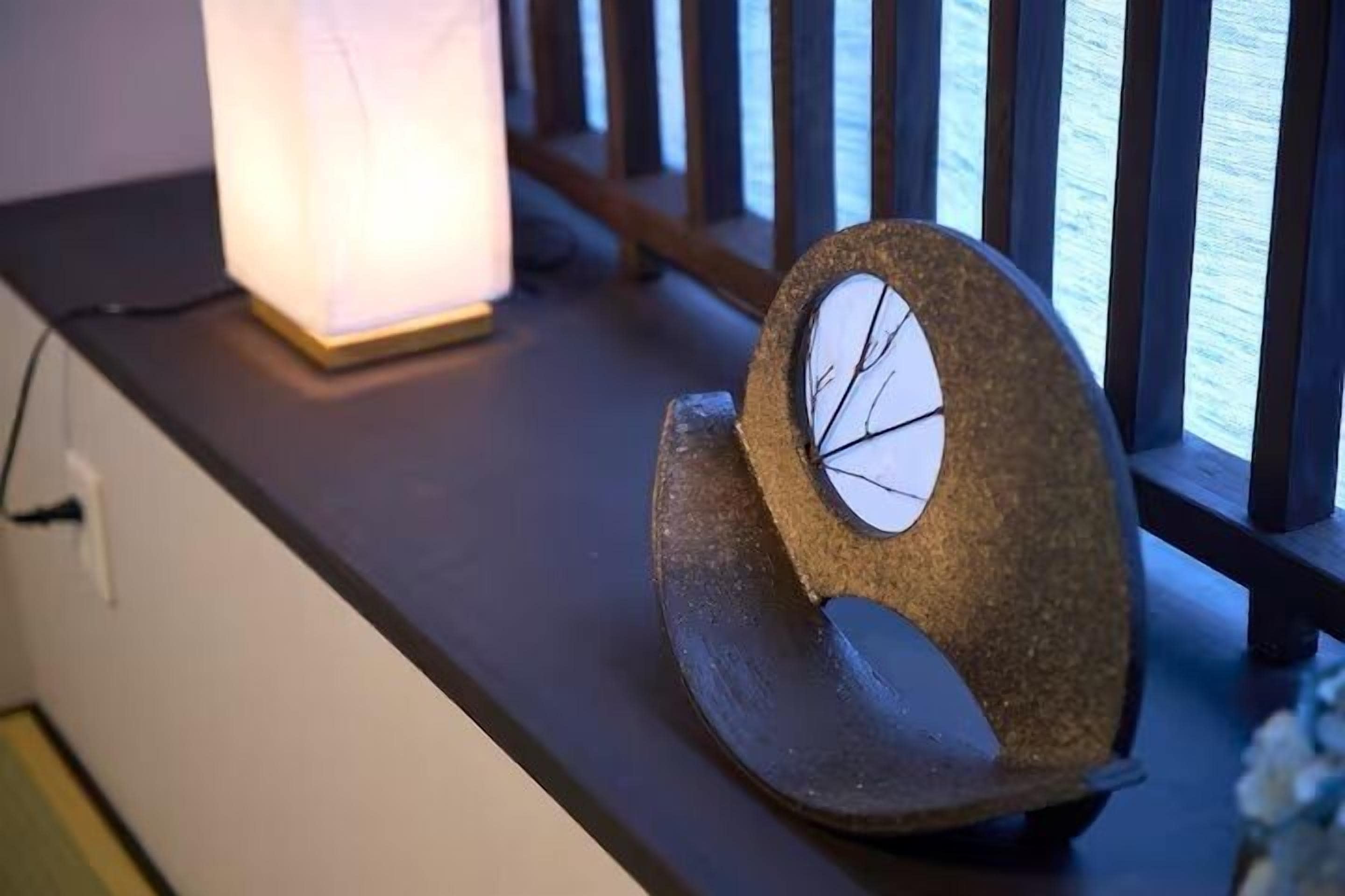 A stone clock with a circular face on a dark wooden surface, next to a lit lamp.