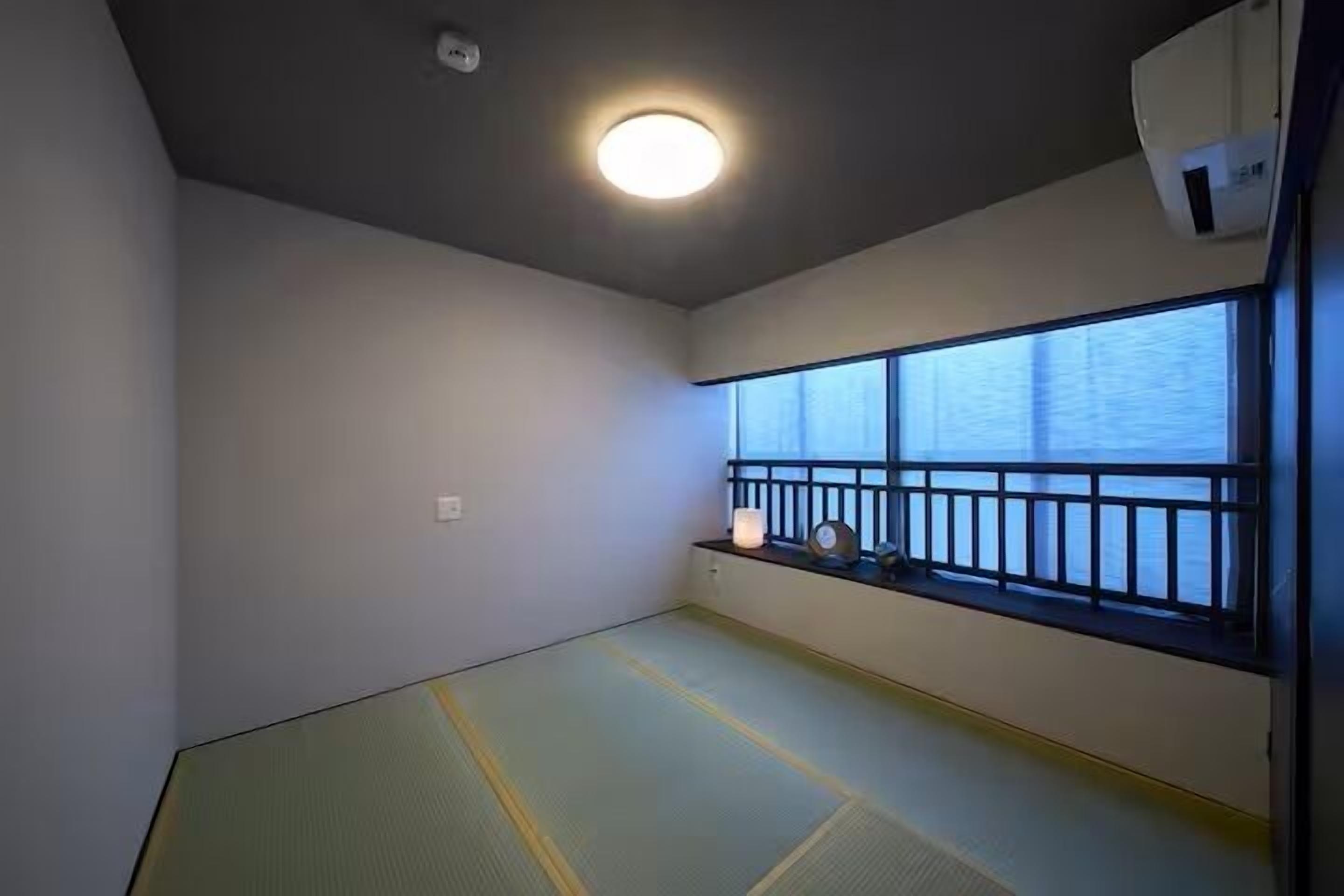 Una habitación con suelo de tatami, un ventanal con barandilla de metal negro y una lámpara en el techo.