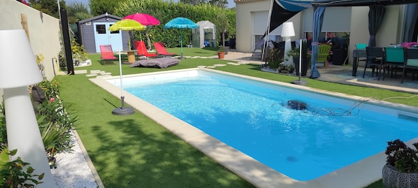 A rectangular swimming pool with a tiled deck, surrounded by a well-maintained lawn and colorful umbrellas.