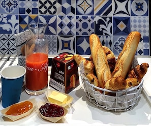 A table with a basket of breadsticks, a glass of tomato juice, a box of butter, and two small containers of spreads.