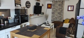Une cuisine moderne équipée d’un four à micro-ondes, d’un four et d’une machine à café. On y trouve une table à manger avec un bol noir et un vase blanc. Un canapé marron et une table basse blanche se trouvent dans l’espace salon.