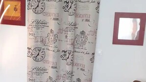 Een bruine bank met een zwart kussen, een decoratief gordijn en een ingelijste foto aan de muur.