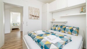 A bedroom with a bed, a nightstand, a wall-mounted shelf, and a door leading to another room.