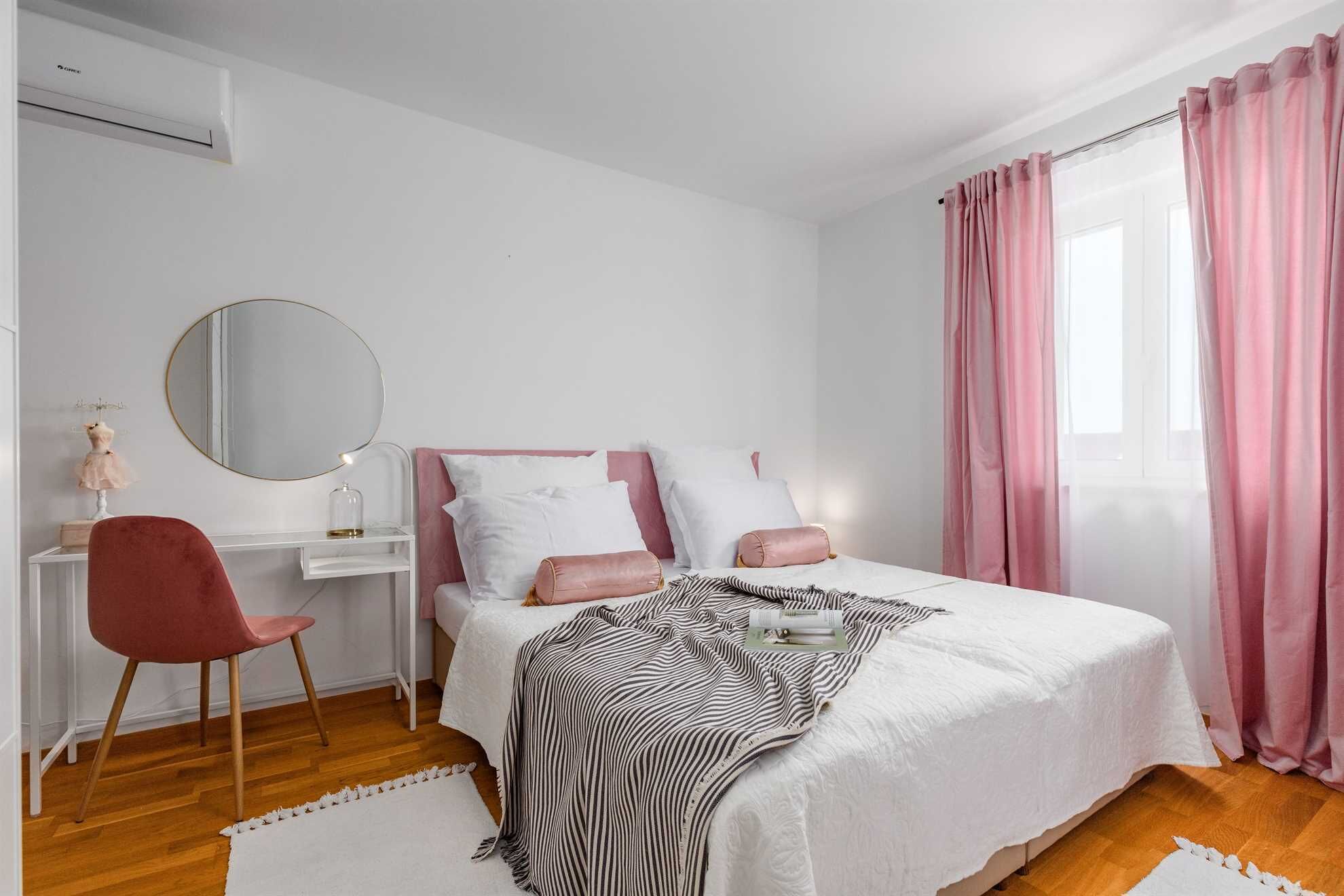 A bedroom with a bed, a desk, a chair, a mirror, and pink curtains.