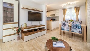 Ein modernes Wohnzimmer mit einem Flachbildfernseher, einer Holzeinbauküche, einem Essbereich mit stuhlbezügen mit Blumenmuster und einer Küche mit weißen Schränken.