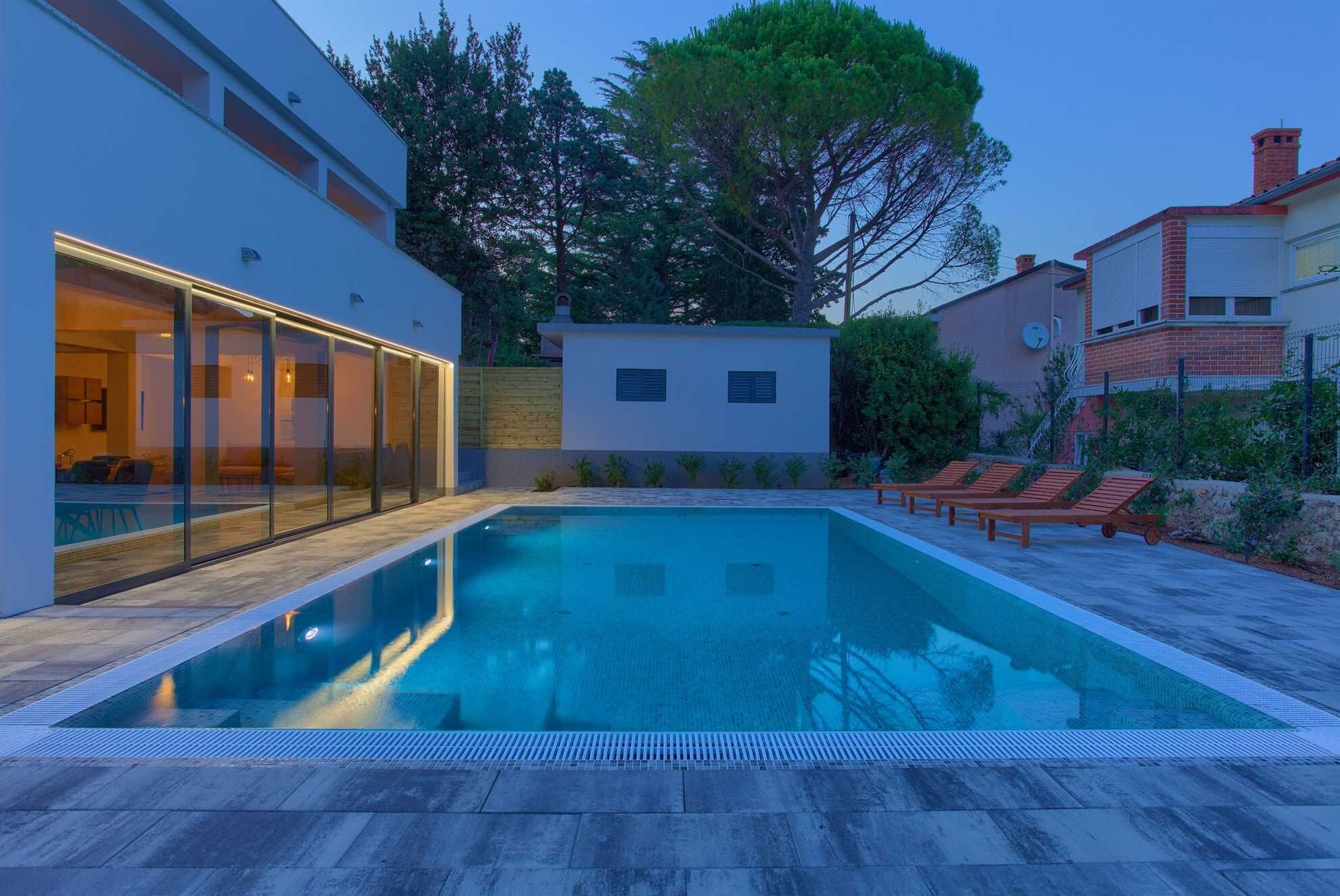 Un espace piscine moderne doté d’un patio vitré, de chaises longues et offrant une vue dégagée sur les bâtiments et les arbres environnants.