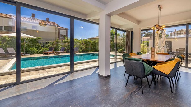 A modern indoor pool area with a dining table and chairs, large windows, and a view of the outside.