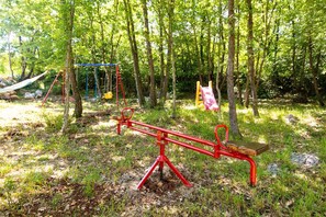 Children can play on the seesaw, swing, and slide in this outdoor playground surrounded by trees.