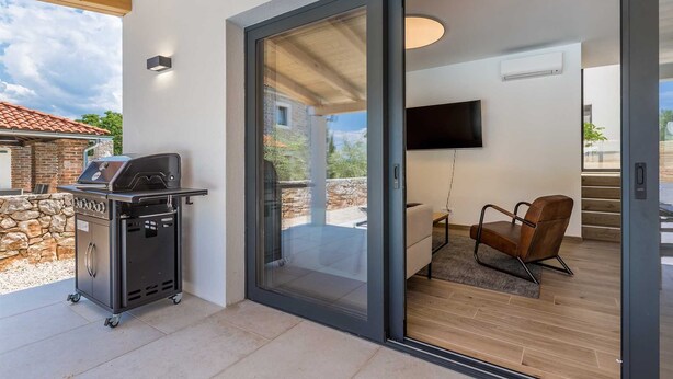 A patio with a barbecue grill, a sliding glass door leading to a room with a TV and a chair.