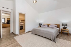 Une chambre à coucher avec un lit, deux tables de chevet, une commode, un plancher en bois et une vue sur la cuisine et la salle de bain, grâce à une porte ouverte.