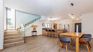 A modern kitchen with a central island, a dining area with wooden tables and chairs, and a staircase leading to an upper floor.