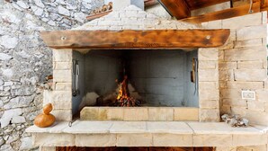 A stone-built outdoor fireplace with a wooden mantel, surrounded by stone walls and a wooden roof structure. - Rustic apartment EVELIN close to the beach (Pinezići)