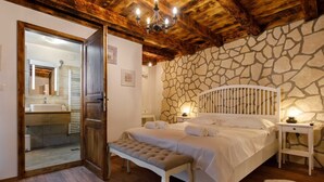 A bedroom with a stone wall, wooden ceiling, a bed with white bedding, a bench, and a nightstand with a lamp. - Rustic apartment EVELIN close to the beach (Pinezići)