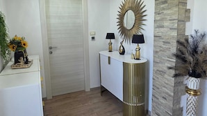 A hallway with a white door, a white console table with two lamps, a gold mirror, and a decorative wall panel.