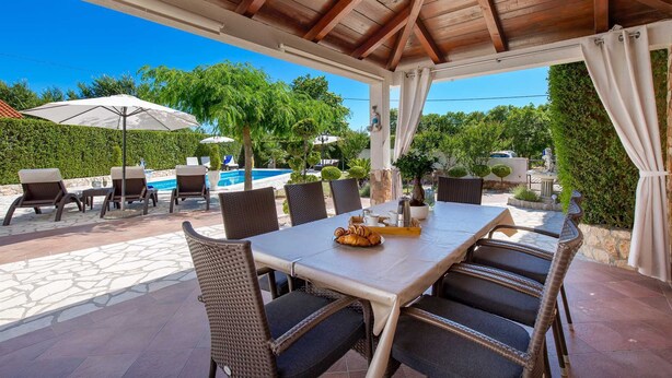 An outdoor patio with a table set for dining, surrounded by wicker chairs, a pool, and greenery.