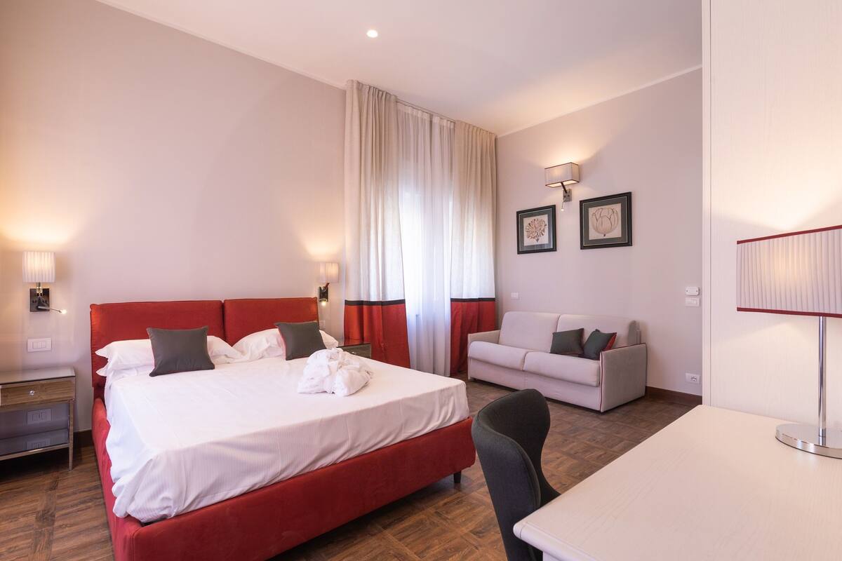 Een hotelkamer met een rood bed, witte gordijnen, een witte bank en een bureau.