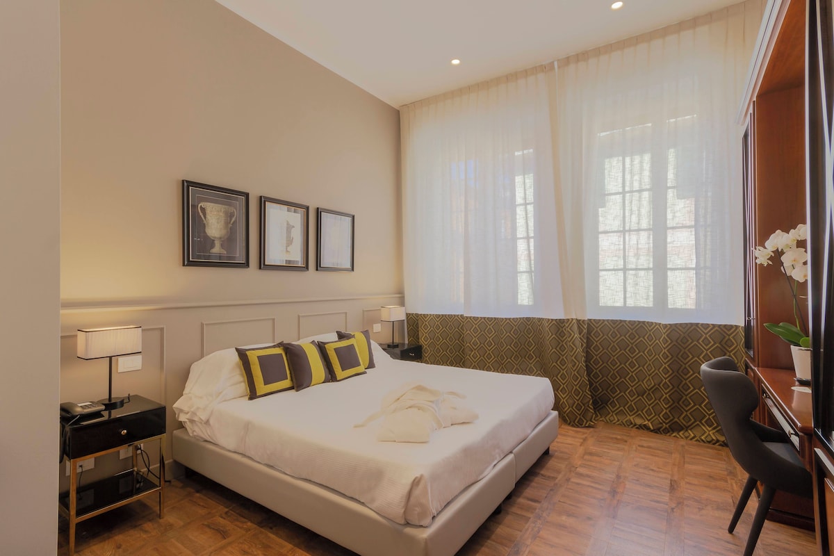 Quarto de hotel bem organizado, com cama, mesas de cabeceira, escrivaninha e janela com cortinas translúcidas.