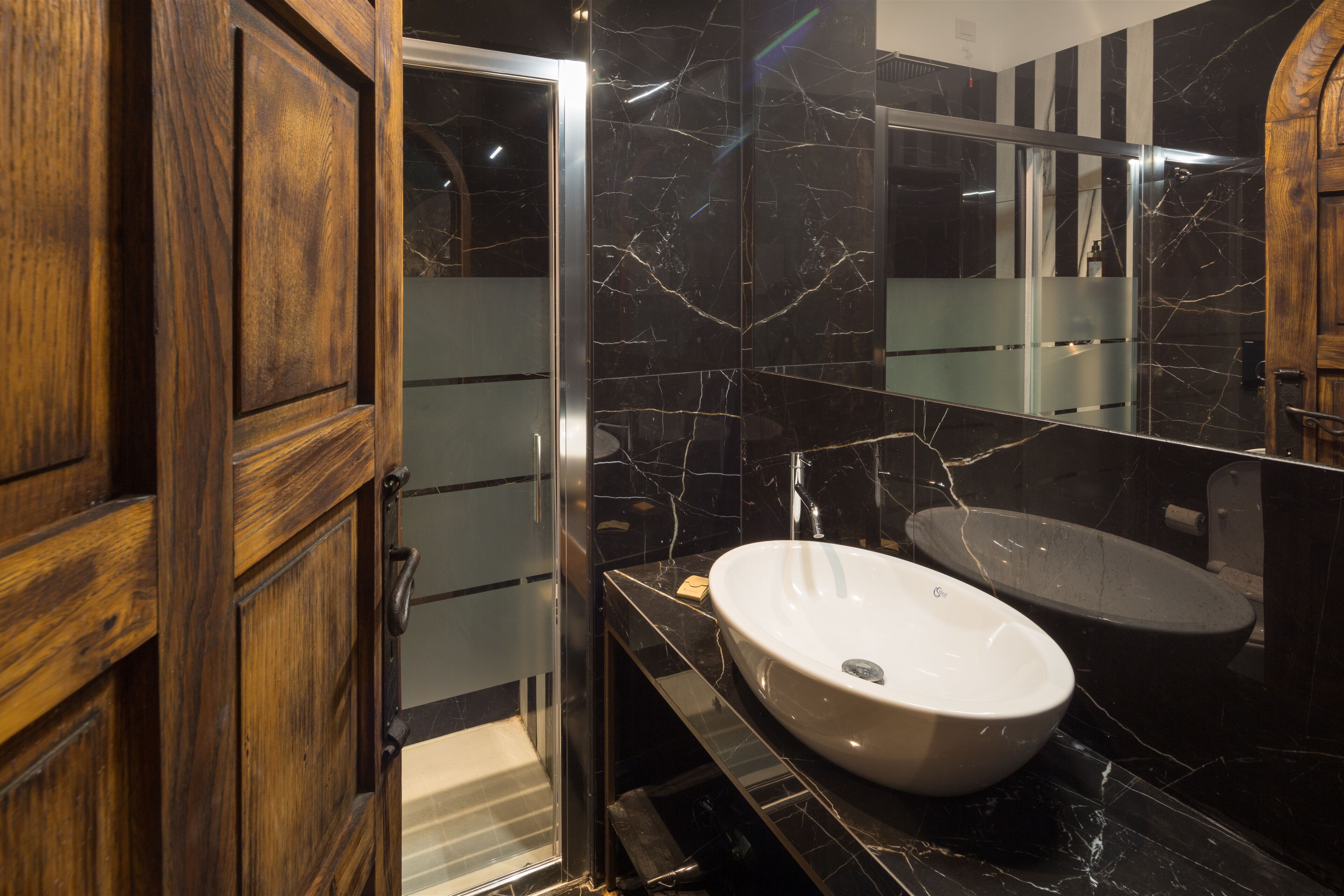A modern bathroom with a wooden door, a glass shower enclosure, a white sink, and a large mirror.