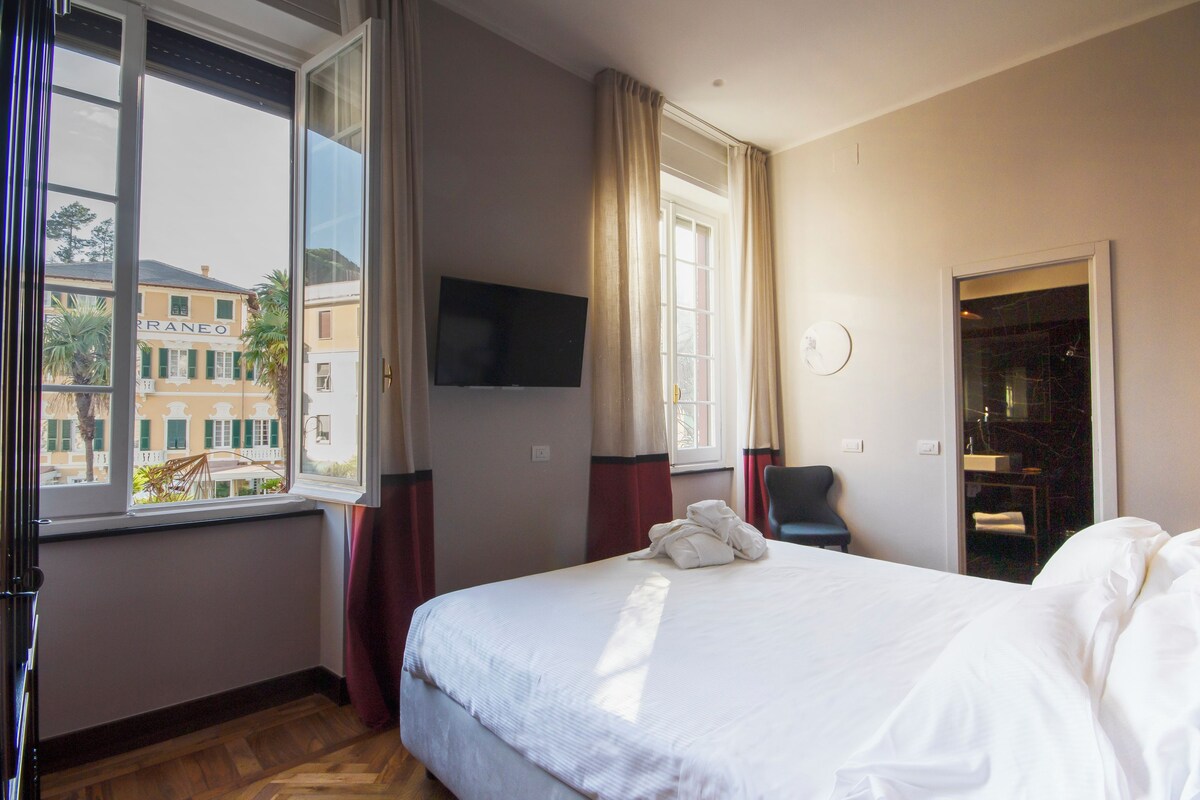 A hotel room with a bed, a TV mounted on the wall, a window with curtains, and a view of buildings outside.