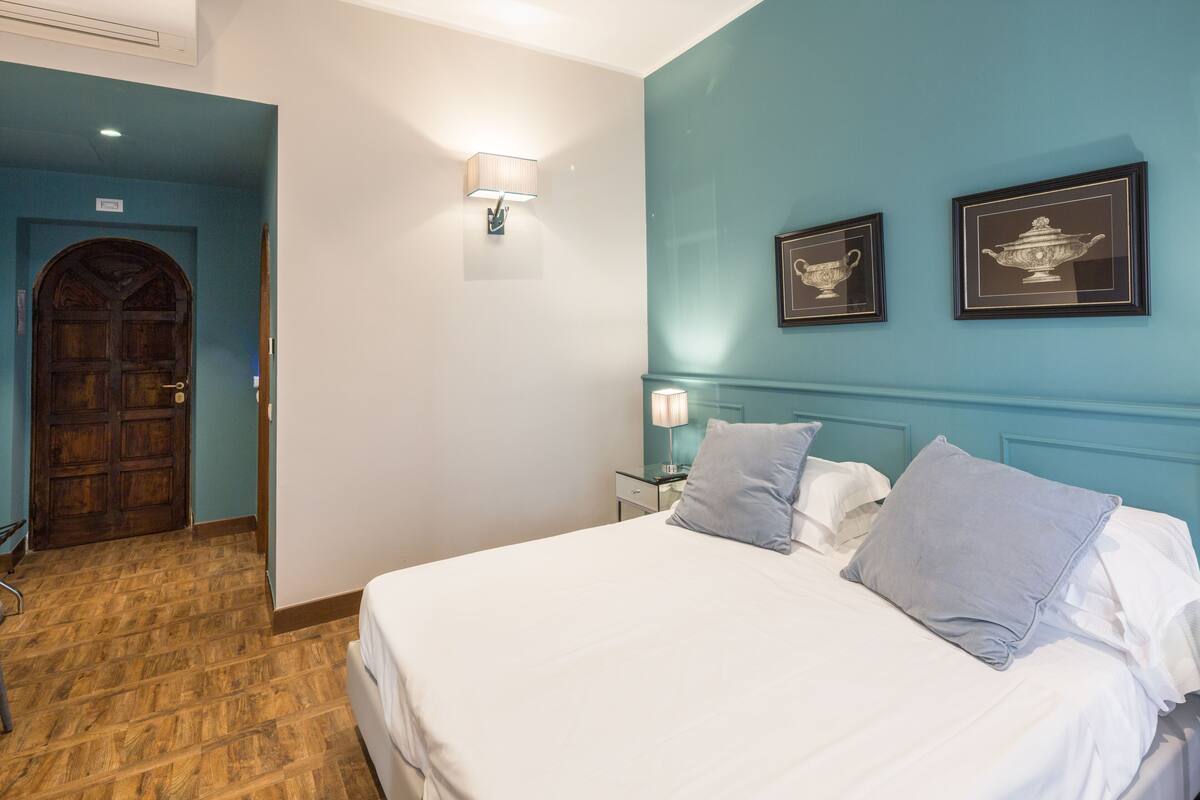 Een hotelkamer met een houten deur, een bed met witte lakens, een nachtkastje met een lamp en twee ingelijste foto's aan de muur.