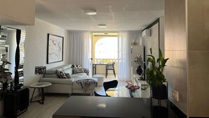 Ein modernes Wohnzimmer mit einer Couch, einem Couchtisch und einem Essbereich. Es gibt ein Fenster mit Vorhängen, eine Pflanze und ein gerahmtes Kunstwerk an der Wand.