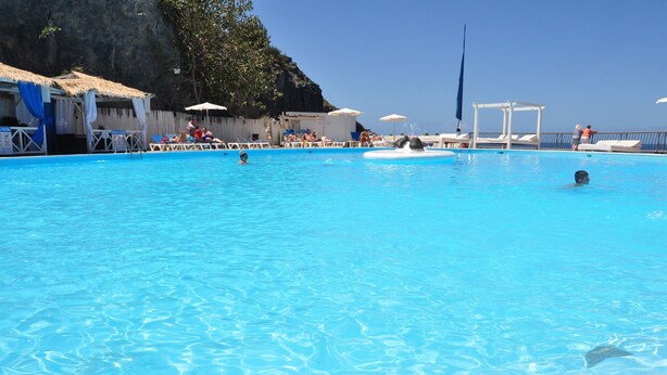 Ein klares, blaues Schwimmbecken mit Liegestühlen und Sonnenschirmen, umgeben von einer felsigen Klippe und blauem Himmel.