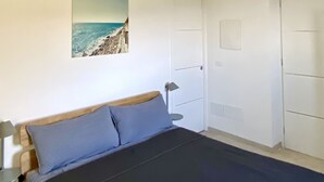 Ett sovrum med en säng, ett nattduksbord, en lampa, en dörr och en tavla som hänger på väggen och föreställer en strandvy.