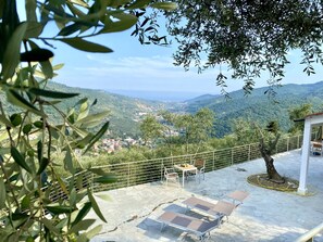 A terrace with lounge chairs and a table, overlooking a scenic landscape with hills and a distant view of the sea. - Dream home with ocean views, a heated pool, and a large terrace in an olive grove (Pontedassio)