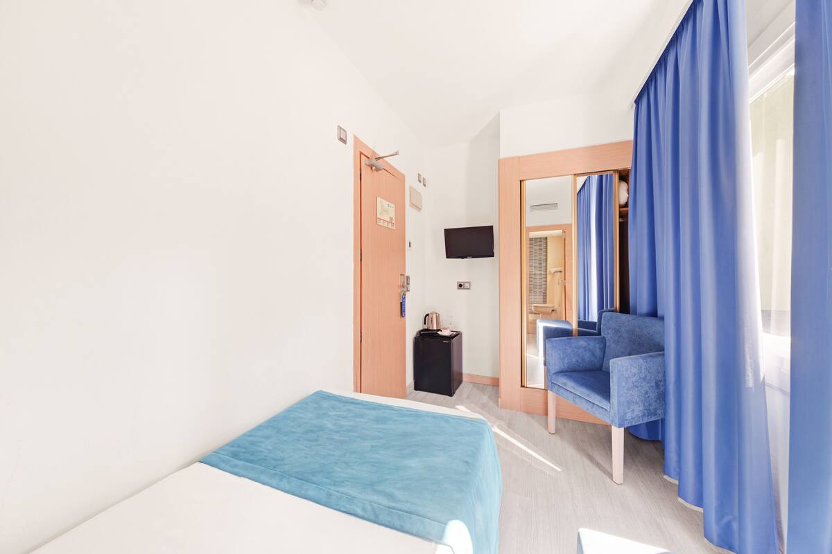 Quarto de hotel com cama, área de estar com cortinas azuis, televisão e pequena mesa com chaleira.