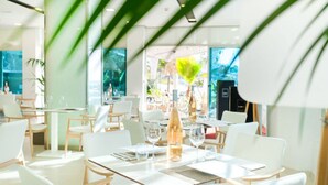 En modern restaurang med vita stolar och bord, stora fönster och utsikt över ett tropiskt landskap.