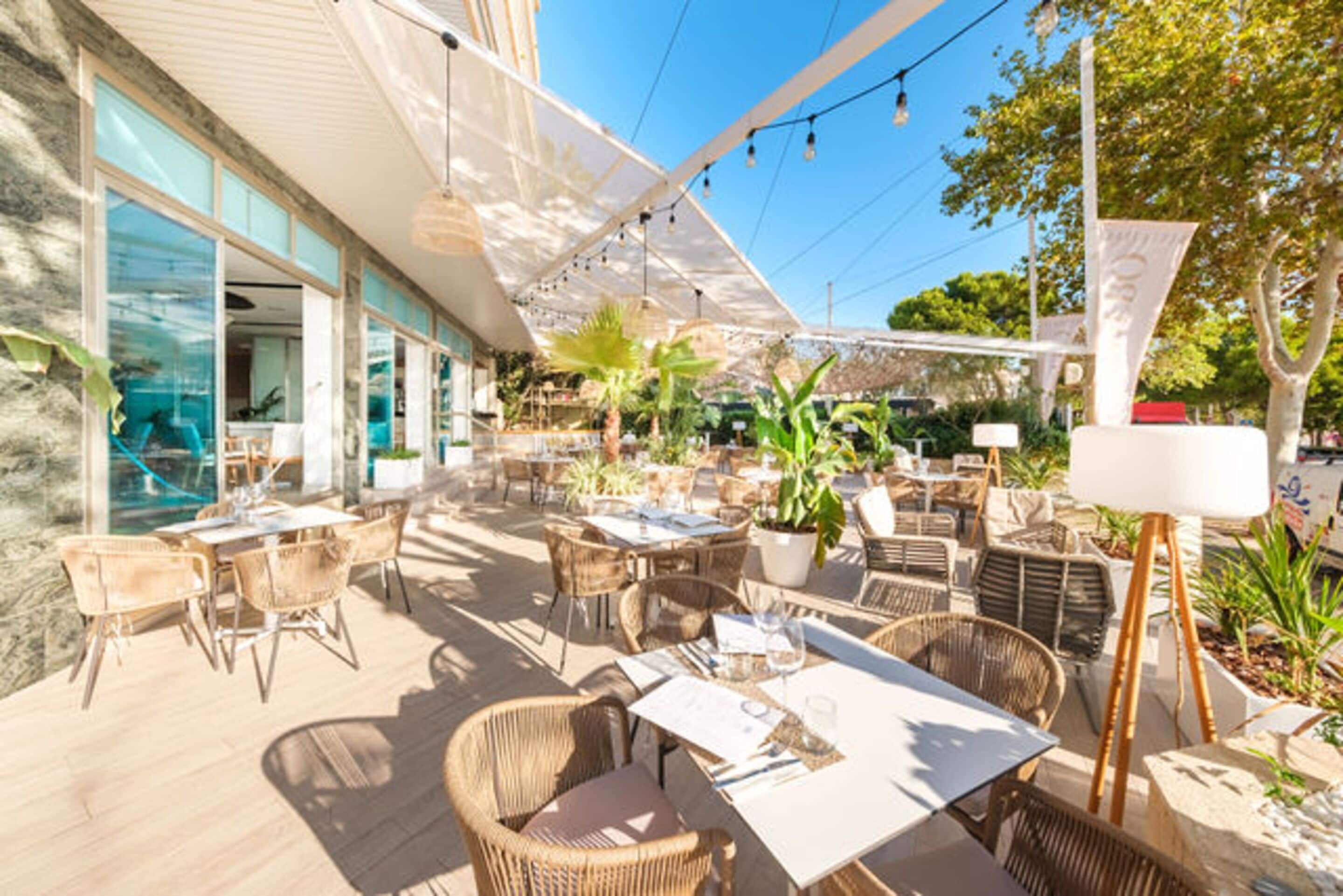 An outdoor patio area with wicker furniture, tables, and chairs, surrounded by potted plants and a large umbrella.