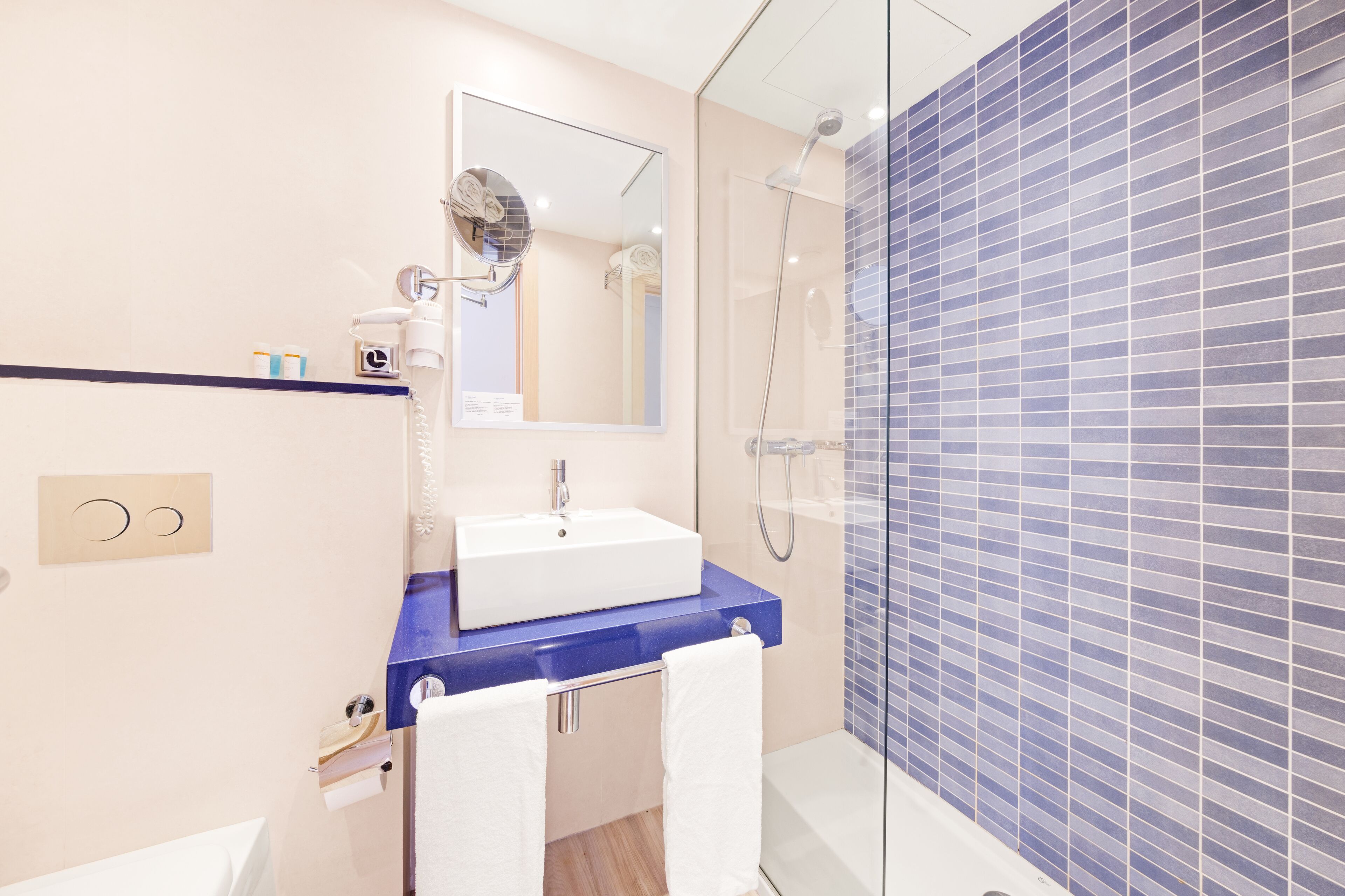 A modern bathroom with a blue countertop, a white sink, a large mirror, and a glass-enclosed shower area.