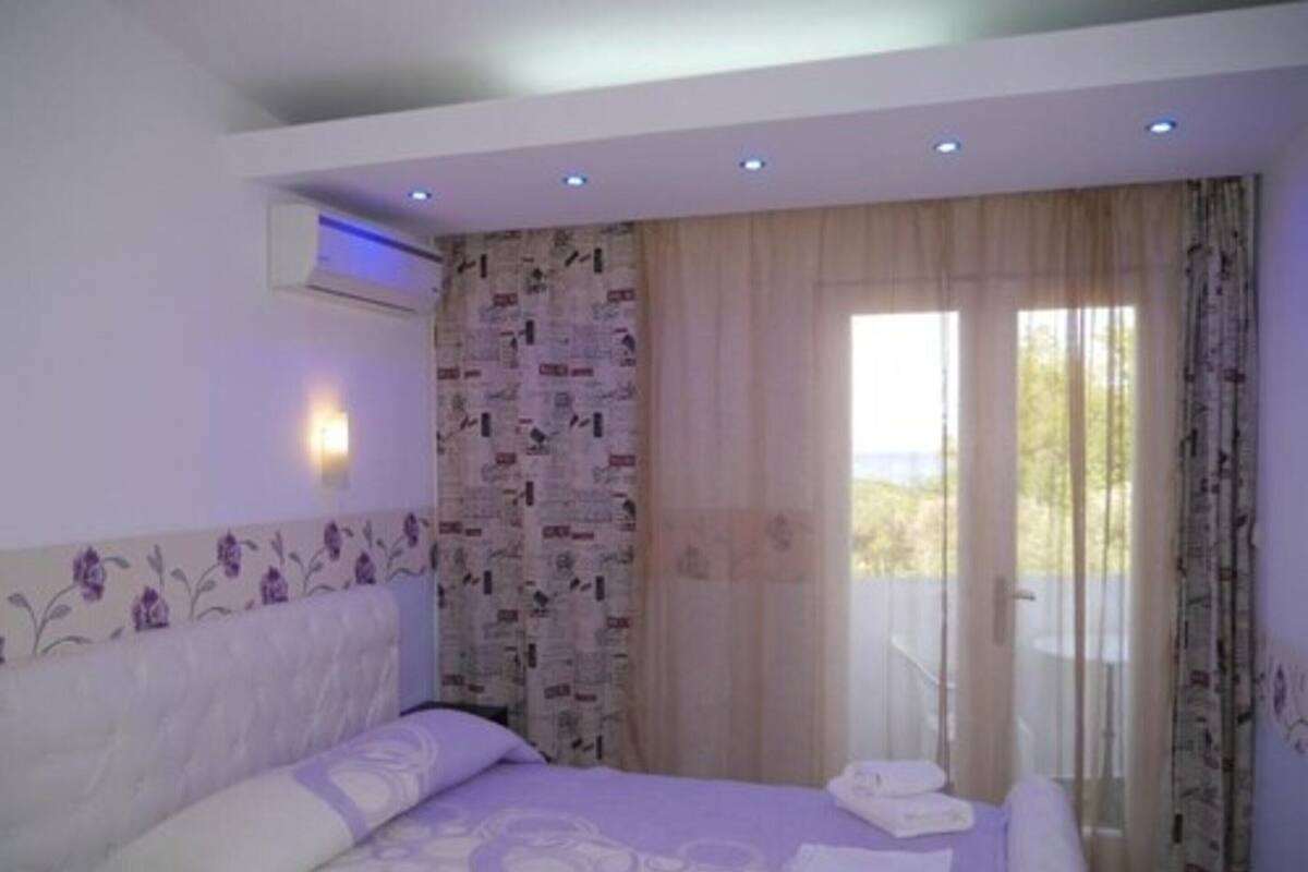 A bedroom with a bed, a window with curtains, a wall-mounted air conditioner, and a balcony with a railing.