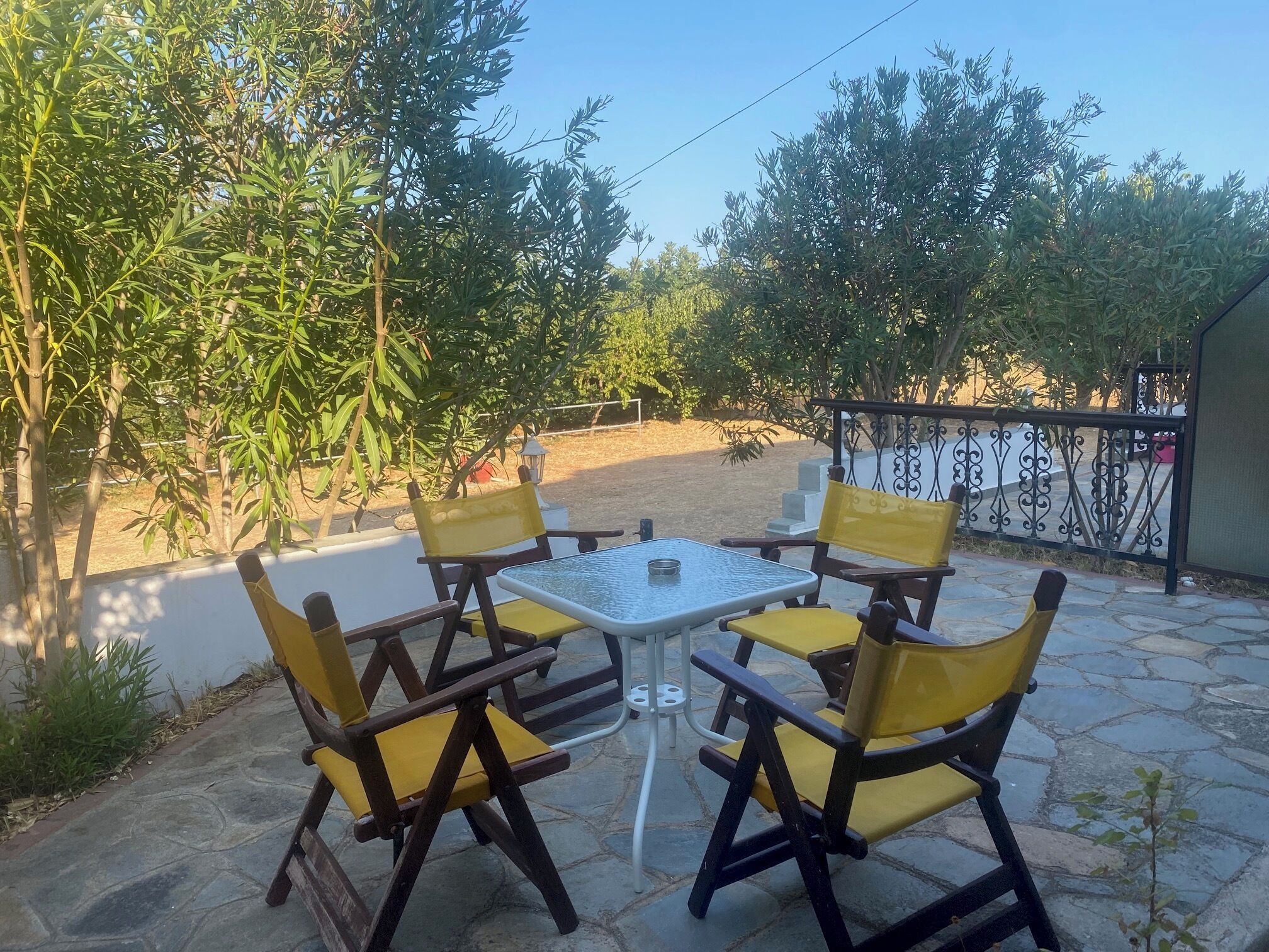 An outdoor patio with a table and four chairs, surrounded by greenery and a fence.