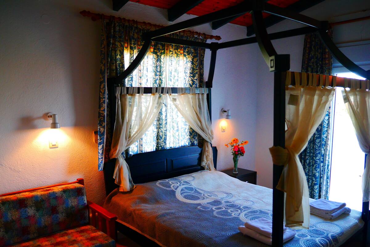 A four-poster bed with a canopy, a bedside table with a vase of flowers, and a chair with a colorful cushion.
