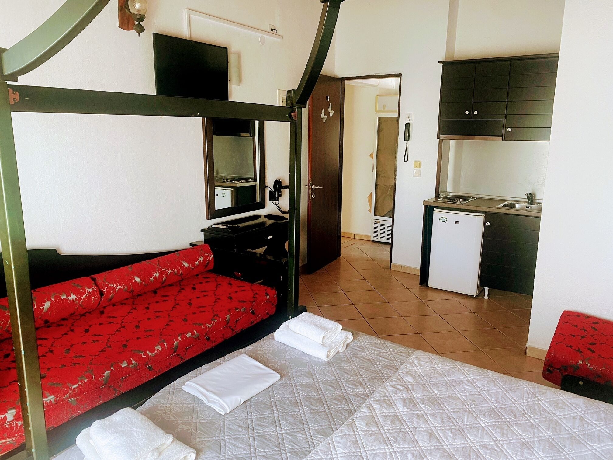 A room with a bed, a red sofa, a small kitchenette, and a door leading to another room.