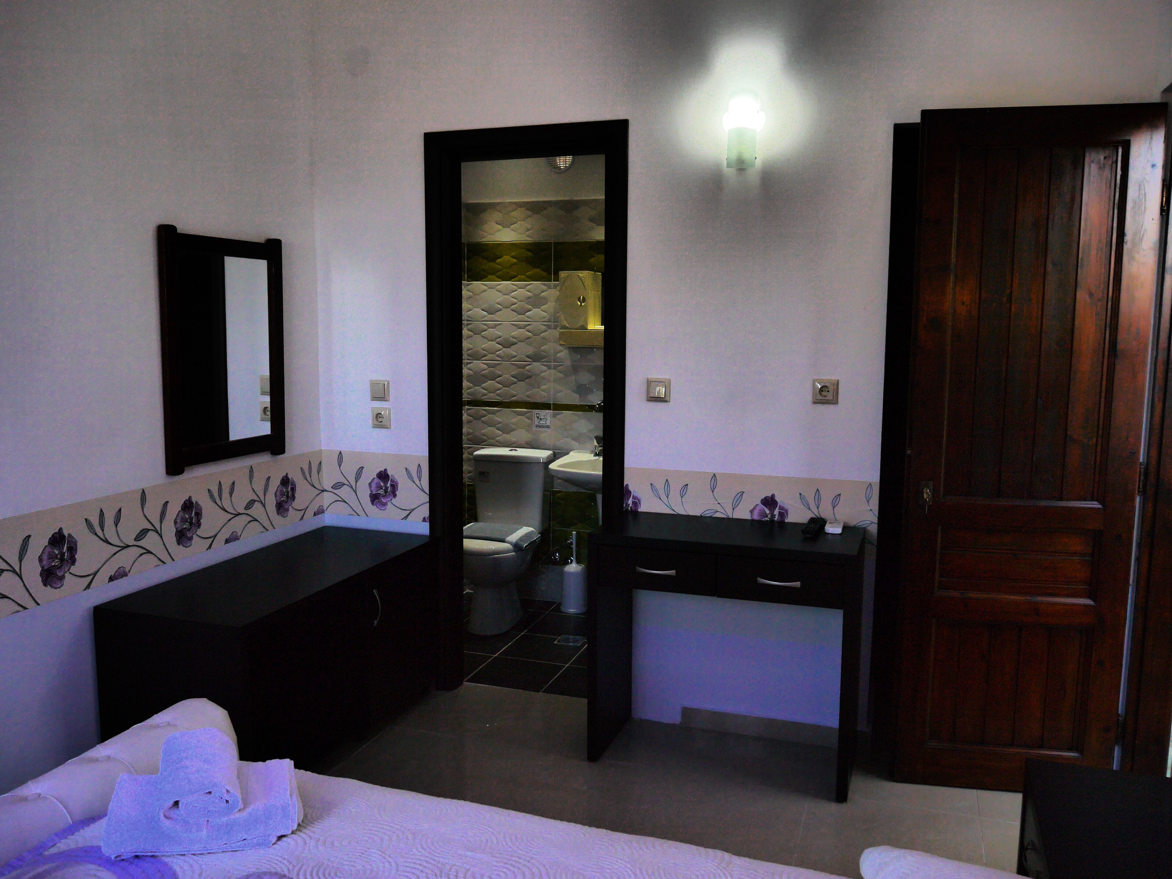Habitación de hotel con una cama, un aparador de madera, un escritorio y un baño visible a través de una puerta abierta.